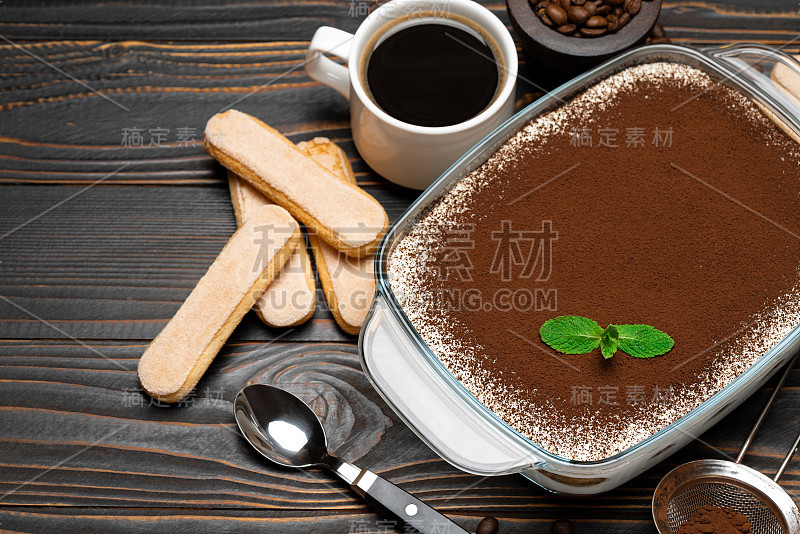 传统的意大利提拉米苏甜点在玻璃烤盘，savoiardi饼干和一杯新鲜的热咖啡在木背景