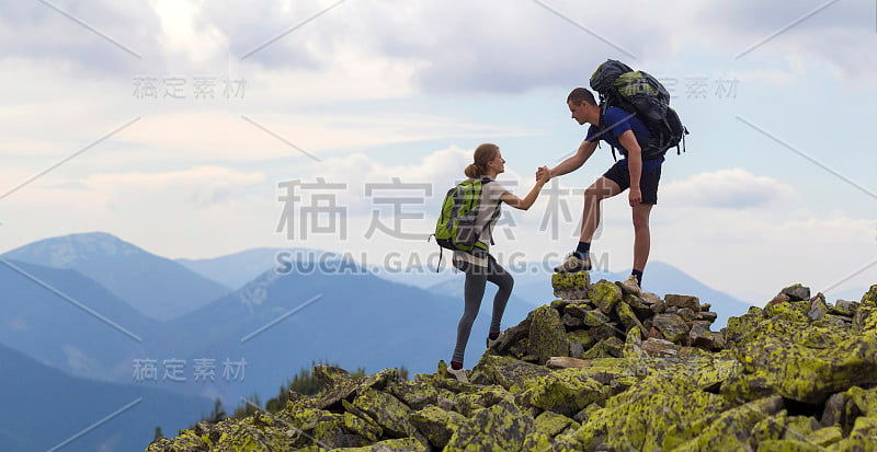 年轻的游客背着背包，运动男孩帮助苗条的女孩在明亮的夏日天空和山脉背景下爬上岩石山顶。旅游、旅游和健康