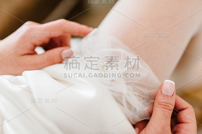 新娘在腿上系吊袜带。美丽的女性腿在婚纱的照片。新娘把长袜穿在脚上。新娘把婚礼吊袜带戴在腿上。前视图。