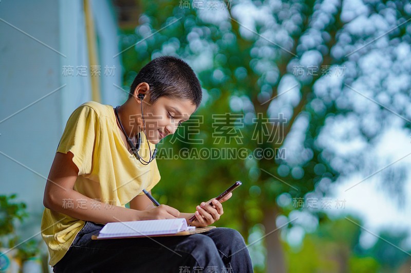 印度一名男孩手持手机，使用手机应用程序进行远程学习，观看在线课程，视频呼叫在应用程序中做笔记，在家学