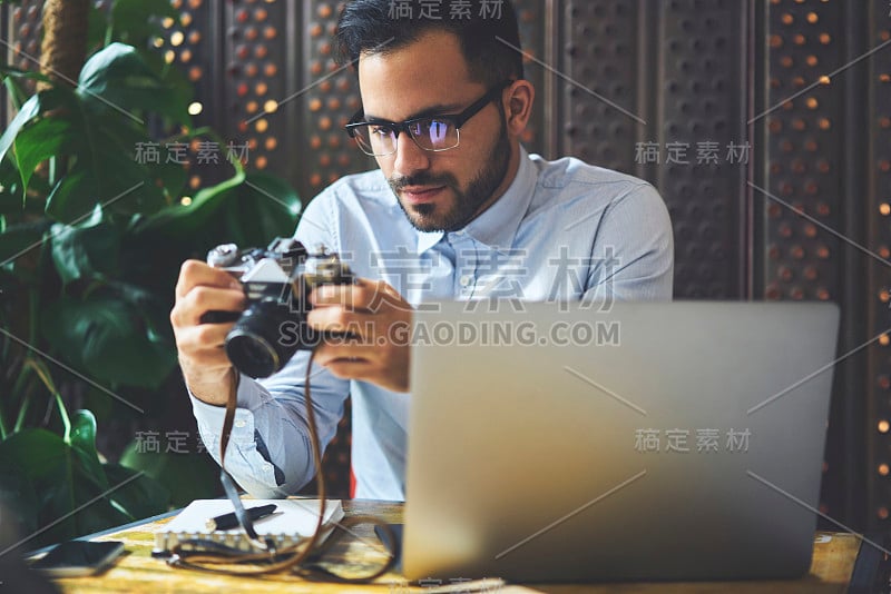 沉思的年轻男性自由职业者坐在咖啡馆的室内，检查老式相机的设置，熟练的摄影师测试设备的可用性，用于拍照