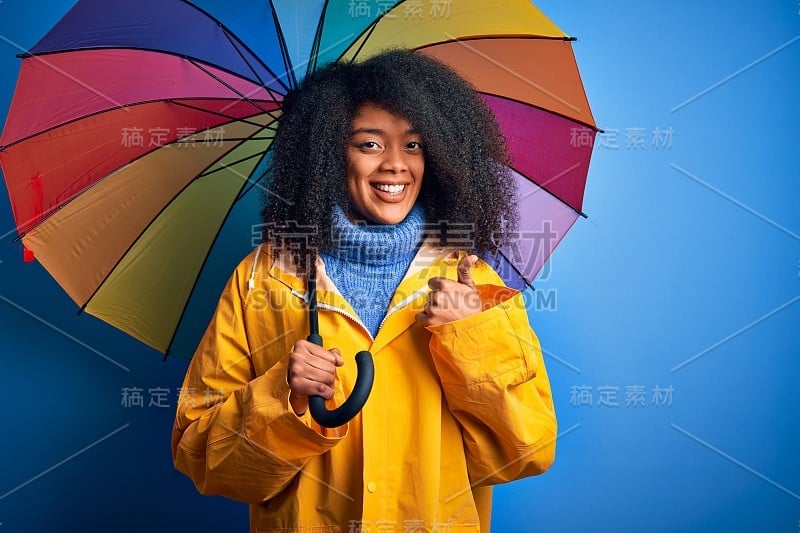 年轻的非洲裔美国妇女与非洲式的头发下彩色伞穿着冬衣下雨高兴与大微笑做ok手势，拇指与手指，优秀的手势