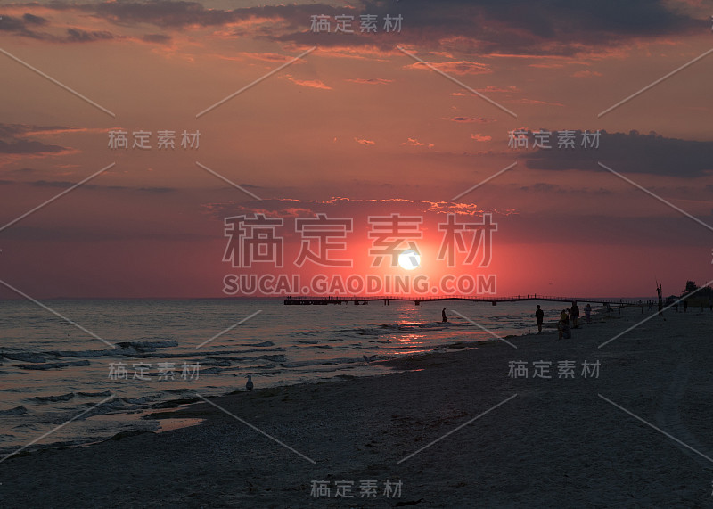 海滩上红色的夕阳。人们在海滩上剪影和海鸥。地平线上的码头