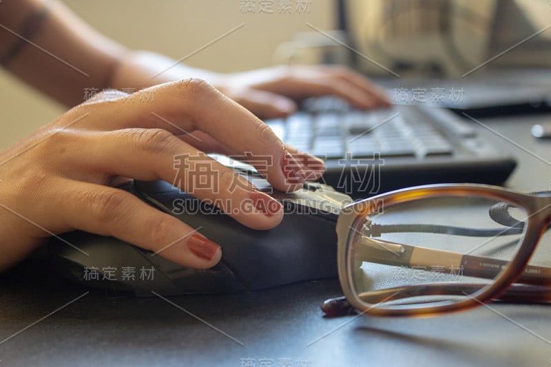 一个女人用她的电脑和眼镜的手的特写，键盘，笔记本电脑和显示器的背景