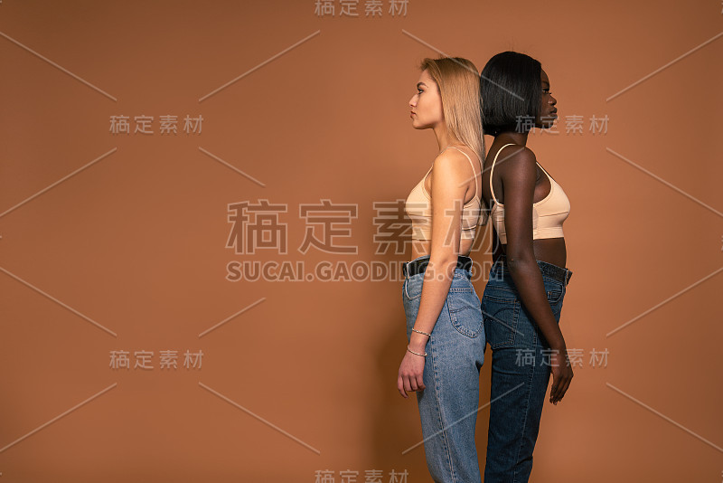 两个国际女孩穿着休闲服背靠背站着，在橙色的背景下孤立地思考