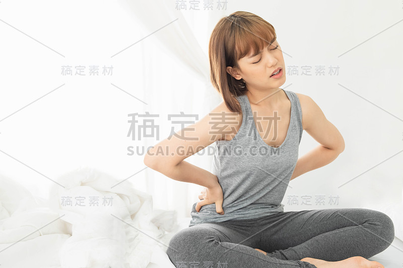 年轻美丽的亚洲女人穿着灰色的衣服，早上醒来在白色的床上，身体疼痛，拥抱的手在腹部蜷缩，腹痛，腹痛，痛