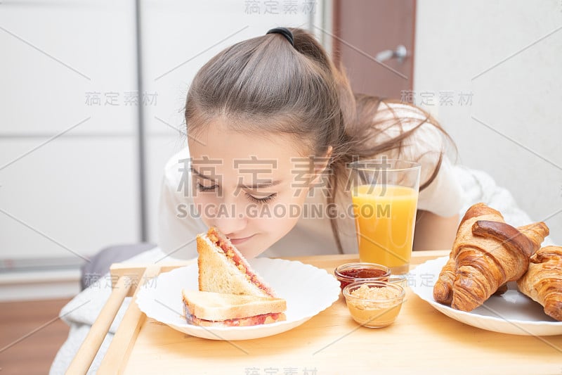 年轻漂亮的女人正在床上享受早餐。羊角面包，橙汁，花生酱和覆盆子果酱三明治。