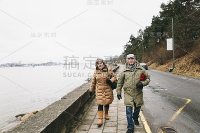 一对老年夫妇——老年男女——穿着暖和的冬装，一起走在海滨附近的人行道上，看着镜头，微笑着