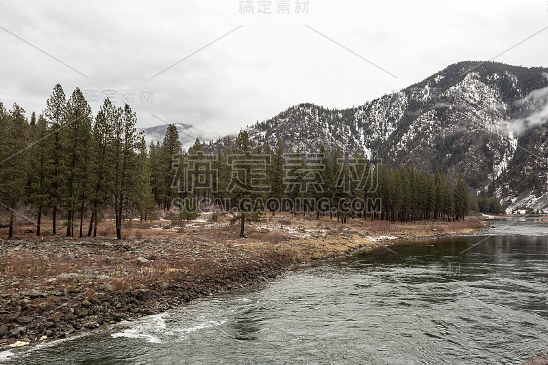 平静的河流，高大的常青树和白雪覆盖的山与雾