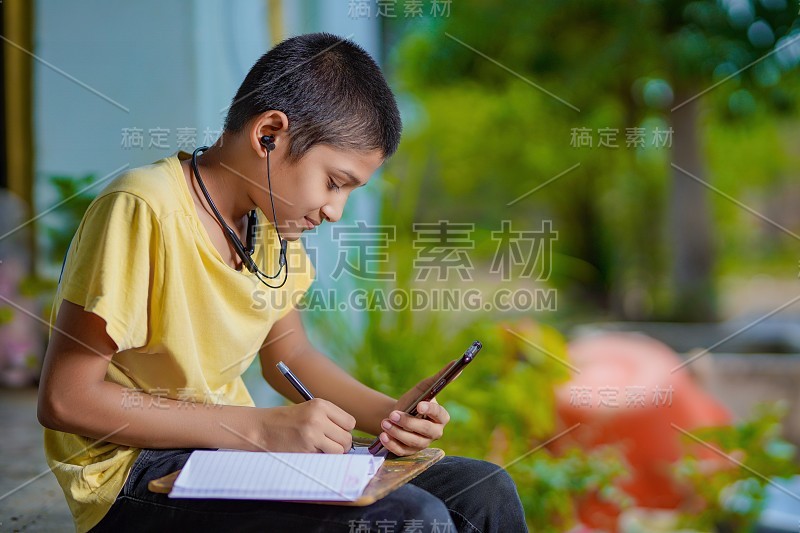 印度一名男孩手持手机，使用手机应用程序进行远程学习，观看在线课程，视频呼叫在应用程序中做笔记，在家学