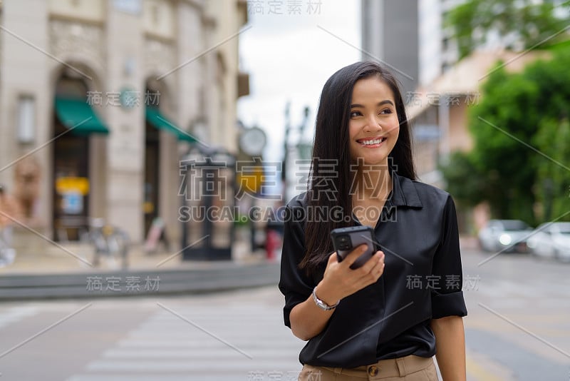 亚洲女商人在户外城市街道上使用手机，同时行走和微笑