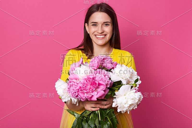 室内拍摄的漂亮的年轻女子拿着美丽的鲜花盛开的白色和粉红色的牡丹花，女士穿着黄色的衣服，摆出孤立的玫瑰