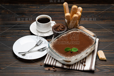 传统的意大利提拉米苏甜点在玻璃烤盘，savoiardi饼干和一杯新鲜的热咖啡在木背景