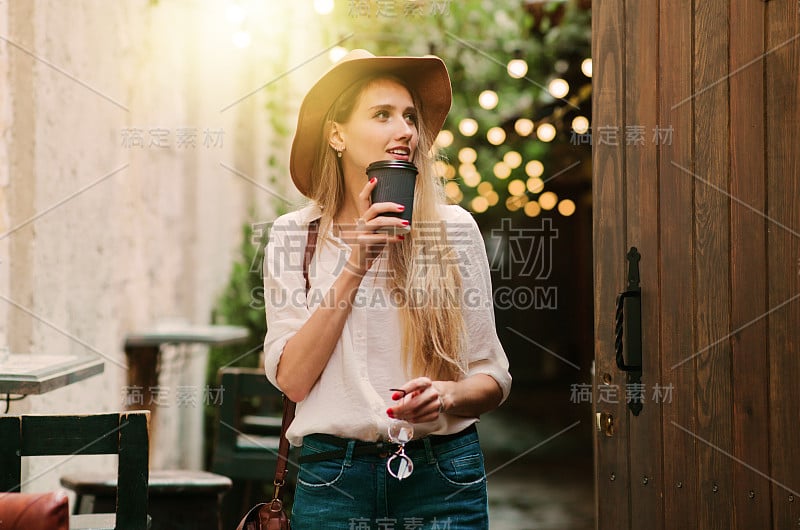 年轻迷人的女人在休闲风格的衣服走在老城的街道上，一杯咖啡在路上