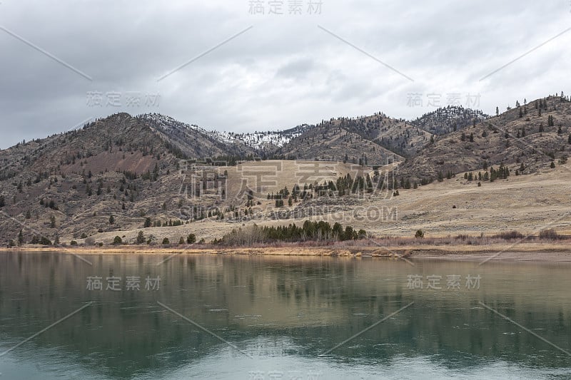 平静的绿色河流前的树覆盖了滚动的山与雪