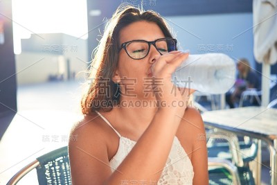 年轻漂亮的女人坐在餐厅里喝着一瓶水享受暑假