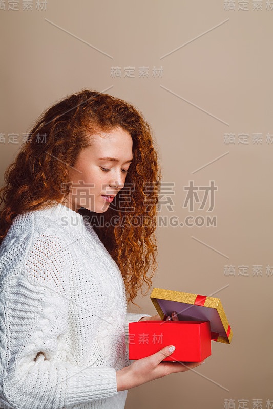 一个穿着白色毛衣，留着长长的卷发的漂亮女孩微笑着打开了一个有金色盖子的红盒子——一个礼物。在摄影棚里