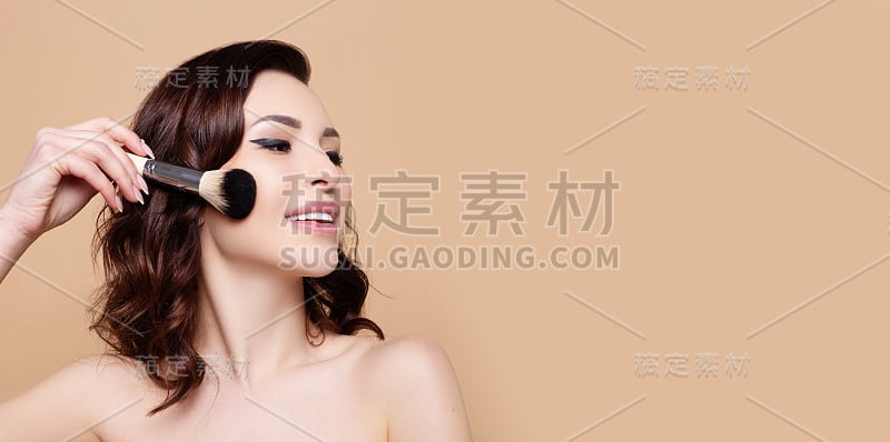 年轻美丽微笑的女人拿着化妆刷在米色的背景。美容院的概念，清新洁净的皮肤，爽肤