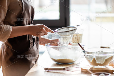 裁剪的年轻女子筛面粉在玻璃碗附近的搅拌器