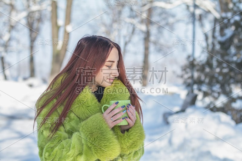 美丽可爱漂亮的红发女青少年在绿色毛皮大衣喝咖啡在阳光明媚的冬季城市公园。