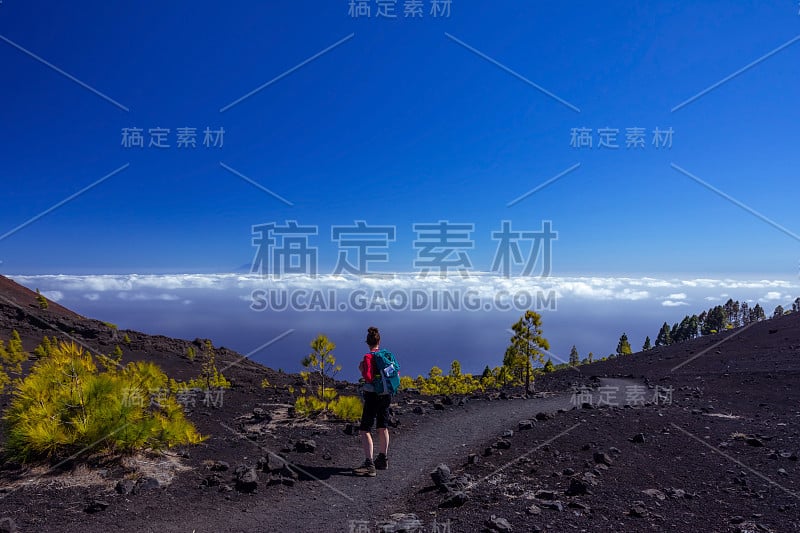 一个背着背包的年轻徒步旅行者，站在一条火山灰小道上，看着地平线上的特内利法岛。