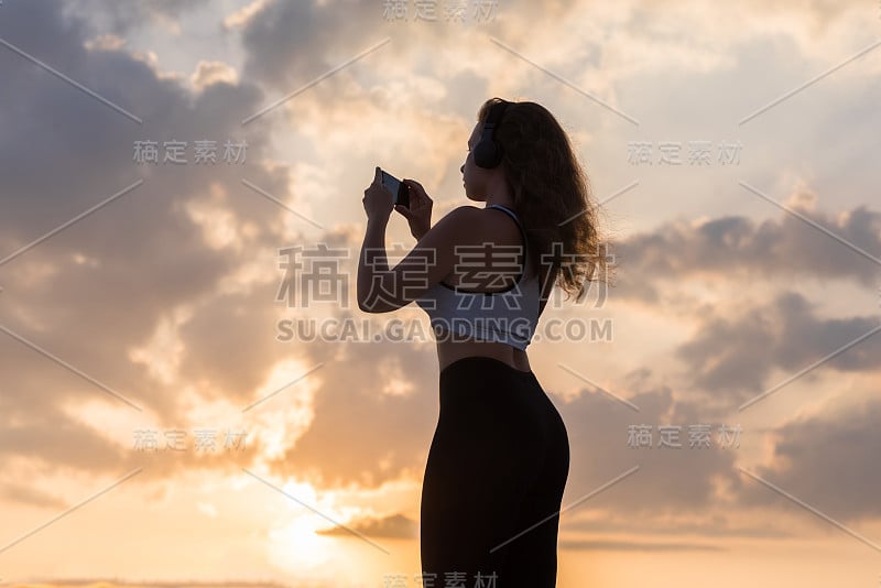 剪影年轻女子在运动服，耳机和手机在日落天空的背景。健康生活方式的概念