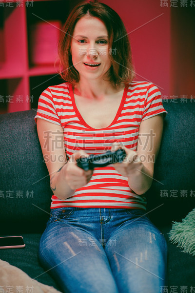 坚定的女孩晚上在客厅玩电子游戏。女玩家坐在沙发上，用无线控制器玩着视频游戏