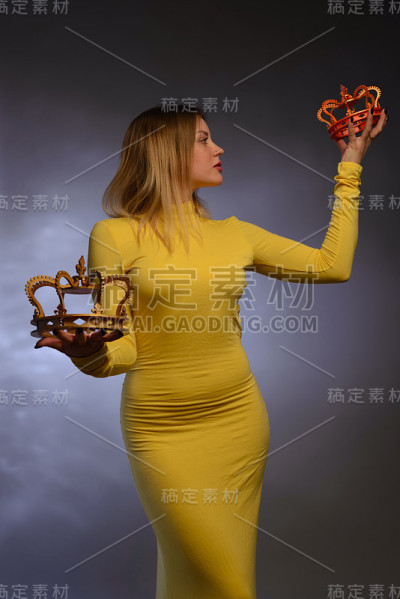美丽的女孩穿着黄色的晚礼服，手里拿着两个皇冠在蓝色的背景