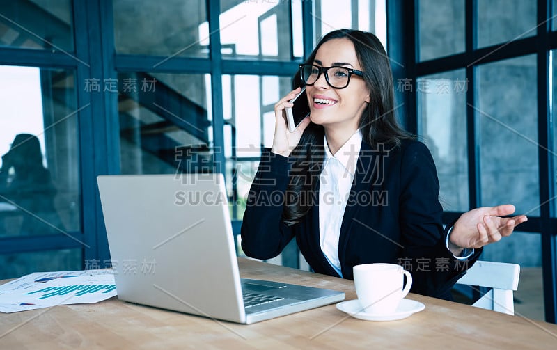 迷人的快乐商务女性穿着全套套装，坐在现代办公室里使用智能手机和笔记本电脑