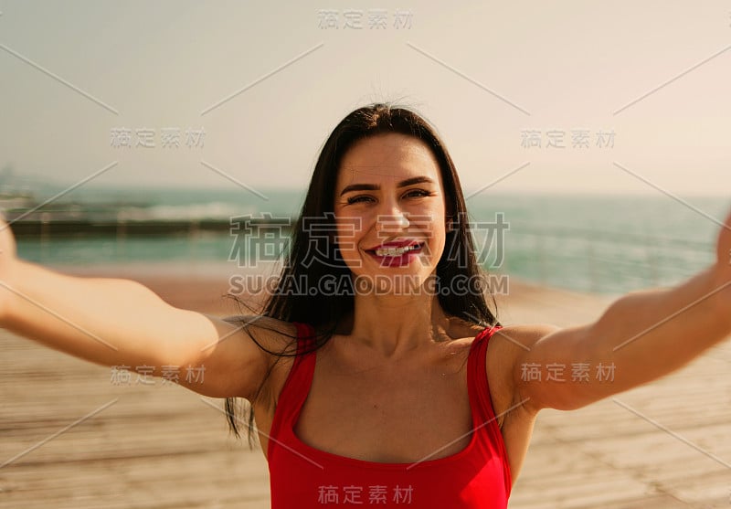 一个年轻的微笑女人在一个红色泳衣在海滩自拍的肖像