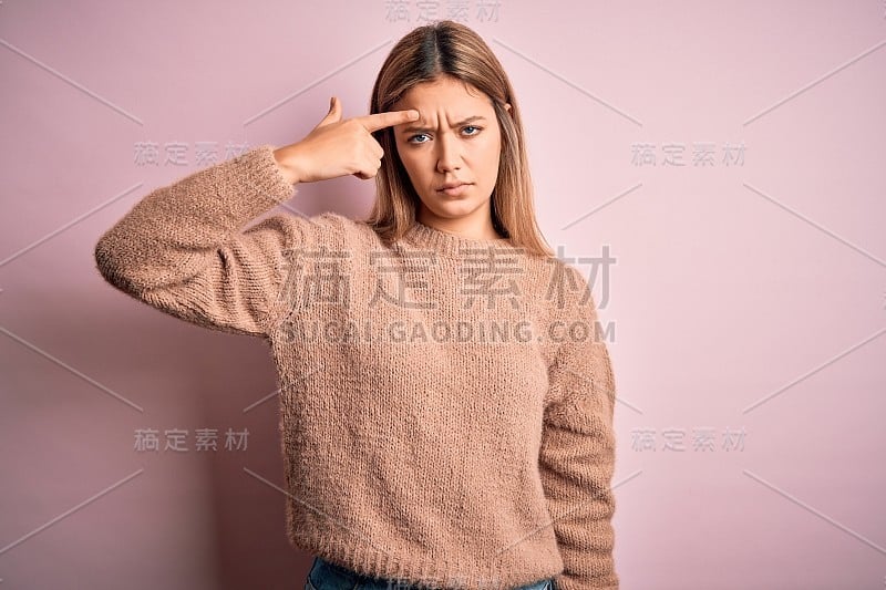 年轻美丽的金发女人穿着冬季羊毛衫在粉红色孤立的背景指向不愉快的丘疹在前额，丑陋的感染黑头。痤疮和皮肤