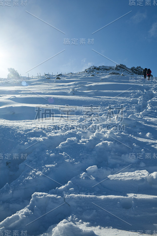 在一个阳光明媚的冬日早晨，两名登山者在雪地上向山顶进发