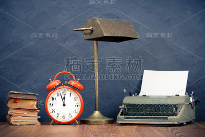 老式打字机与空白纸，橙色闹钟，旧台灯，老书在木制桌子前混凝土黑色墙背景。复古风格滤镜照片