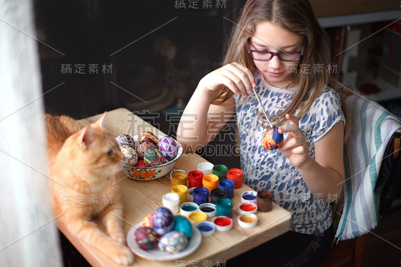 十几岁的女孩画复活节彩蛋，她的姜黄色的猫躺在桌子上。