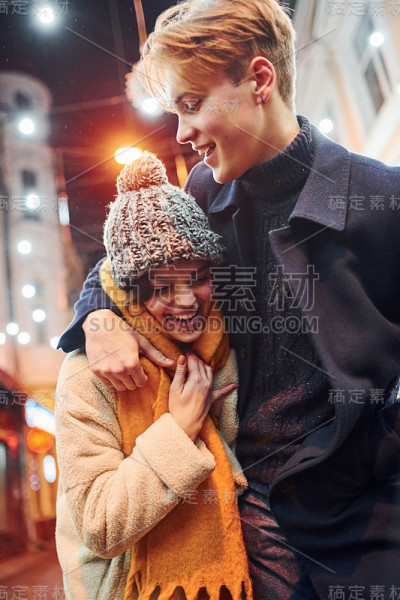 积极的年轻夫妇在温暖的衣服拥抱对方在圣诞装饰的街道