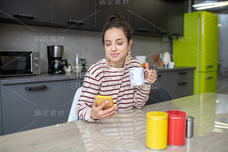 一个年轻女人坐在厨房里，拿着杯子，查看手机