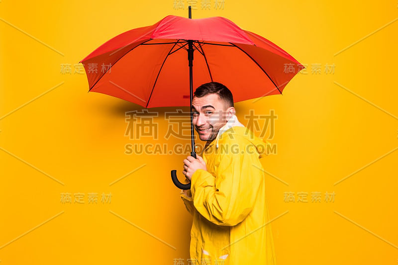 侧面的年轻英俊的胡子男人在黄色雨衣与红色伞思考孤立在橙色背景