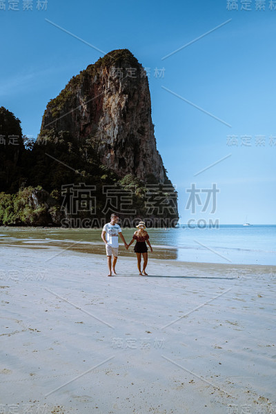 泰国甲米海滩，一对情侣在海滩上散步，热带悬崖和长尾船在甲米海滩岛的背景中