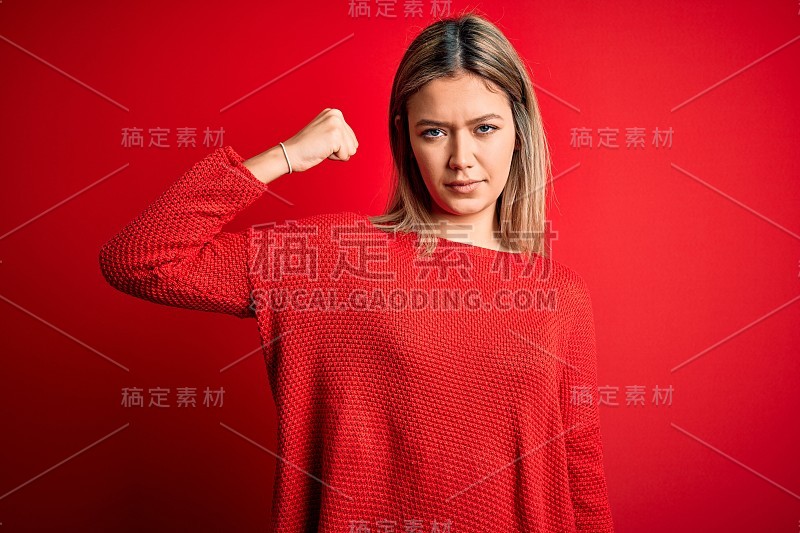 年轻美丽的金发女子穿着休闲毛衣在红色孤立的背景强壮的人显示手臂肌肉，自信和自豪的力量