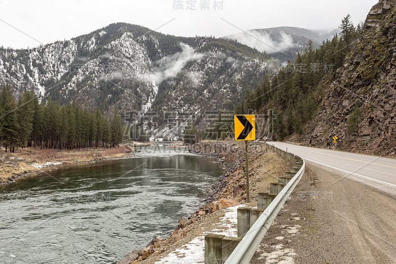 路标指向空旷的高速公路周围的河流和白雪覆盖的山在雾