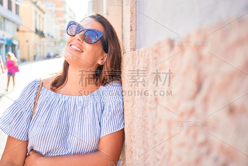 在一个阳光明媚的夏日，年轻美丽的女子靠在城市街道的墙上，快乐地微笑着