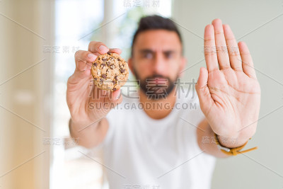 英俊的西班牙男子吃巧克力片饼干与开放的手做停止标志与严肃和自信的表情，防御手势