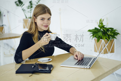 微笑的广告文案手拿一杯卡布奇诺看着镜头，同时在现代笔记本电脑的键盘上打字。美丽的自由职业者坐在木桌与