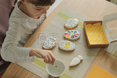 一个扎着辫子的小女孩坐在厨房的桌前，用画笔在复活节饼干上涂颜料