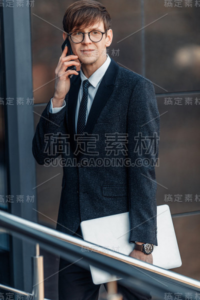 戴着眼镜的时尚商人在商务中心的玻璃大楼附近打电话