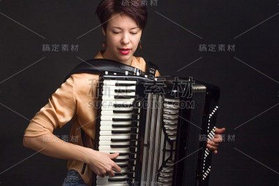 一个穿着金色衬衫、留着短发的黑发女人站在工作室的黑暗背景上。拉手风琴