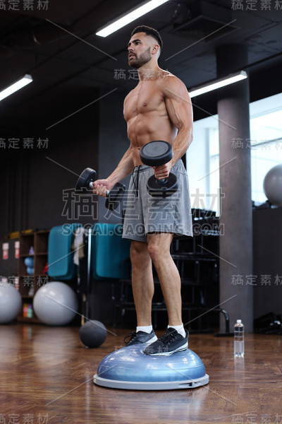 健美和肌肉发达的人在健身房用哑铃在健身球上锻炼。