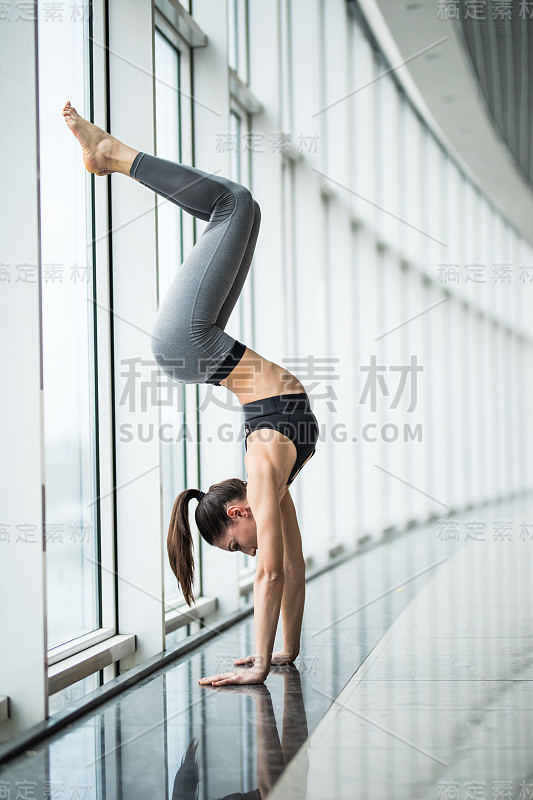 年轻女子练习瑜伽站在手倒立练习，平查Mayurasana姿势与弯曲的腿变化，工作，对全景窗口