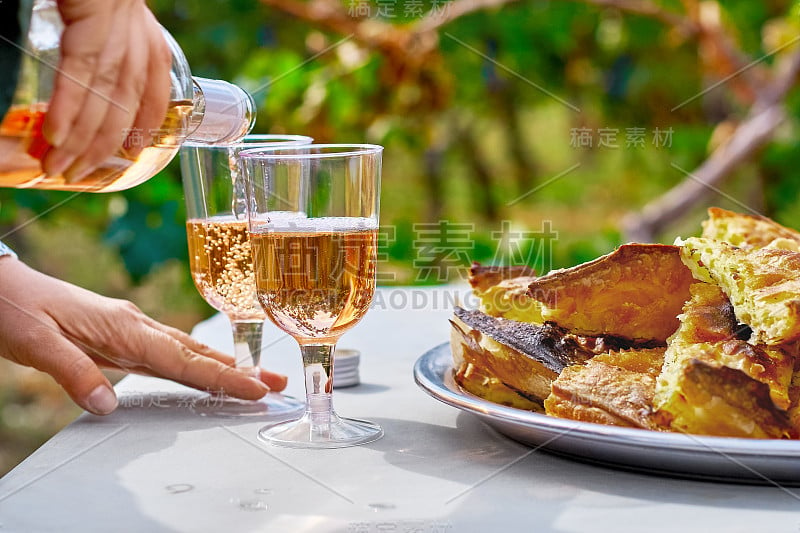 以葡萄园为背景，女人们用手将清爽的粉红葡萄酒倒进玻璃杯，蛋糕摆在桌上。酿酒的概念。