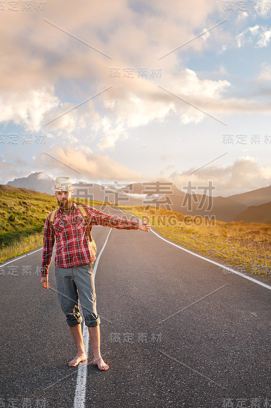 有型的赤着脚、留着胡子、头戴帽子、背着背包的搭车男士在日落时分沿着郊区的柏油路走着，停了下来